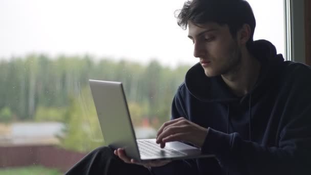 Um jovem está digitando em um laptop sentado na janela contra o pano de fundo de uma paisagem rural pitoresca. O tipo trabalha num portátil na aldeia.. — Vídeo de Stock