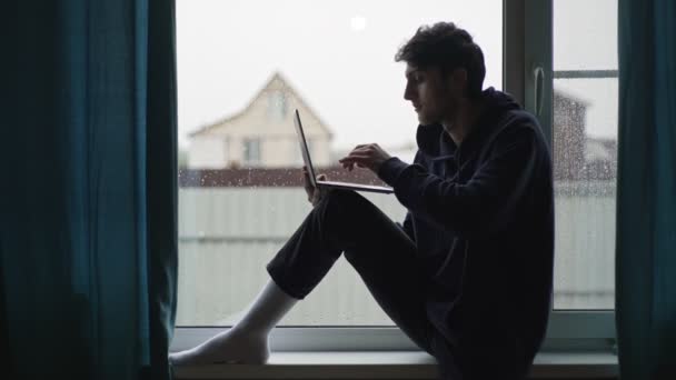 Um jovem está digitando em um laptop sentado na janela contra o pano de fundo de uma paisagem rural pitoresca. O tipo trabalha num portátil na aldeia.. — Vídeo de Stock