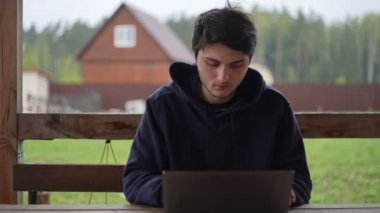 Genç bir adam bir terasta otururken dizüstü bilgisayarında resim gibi kırsal bir arazinin arka planına karşı yazı yazıyor. Adam köyde bir dizüstü bilgisayarda çalışıyor..