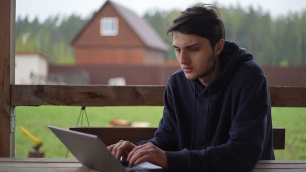 Молодой человек печатает на ноутбуке, сидя на террасе на фоне живописного сельского пейзажа. Парень работает на ноутбуке в деревне.. — стоковое видео