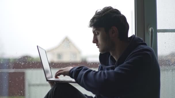 Um jovem está digitando em um laptop sentado na janela contra o pano de fundo de uma paisagem rural pitoresca. O tipo trabalha num portátil na aldeia.. — Vídeo de Stock