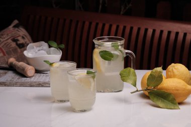Sürahi ve masada iki bardak buz gibi limonata ve limon. Beyaz mermer bir masada gözlüklerle naneli limonata ve arkada ahşap bir bank..