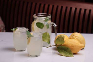 Sürahi ve masada iki bardak buz gibi limonata ve limon. Beyaz mermer bir masada gözlüklerle naneli limonata ve arkada ahşap bir bank..