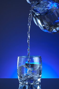 Water poured into a glass, splash, blue background, refreshing, freshness and health. Water bottle, water pitcher, blue liquid, ice, drops, motion, wave, splash, transparency blue liquid on water bottle or pitcher, ice, drops. Gradient background.