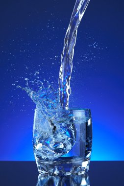 Water poured into a glass, splash, blue background, refreshing, freshness and health. Water bottle, water pitcher, blue liquid, ice, drops, motion, wave, splash, transparency blue liquid on water bottle or pitcher, ice, drops. Gradient background.