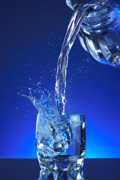 Water poured into a glass, splash, blue background, refreshing, freshness and health. Water bottle, water pitcher, blue liquid, ice, drops, motion, wave, splash, transparency blue liquid on water bottle or pitcher, ice, drops. Gradient background. Stockbild