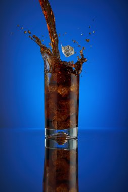 Splash cam kola meşrubat mavi bir arka plan üzerinde yenileniyor. Soğuk sıvı İçecek coca buz ile soğuk bir bardak içine. Restoran ve bar teşvik için içecek. Köpüklü izole Closeup dizayn kahverengi likör.