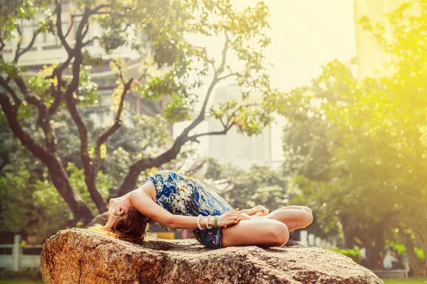 Mooi jong meisje beoefenen van yoga op een rots in een park. In het Park, zonnig weer. Op de achtergrond van een mooie boom en tempel. — Stockfoto