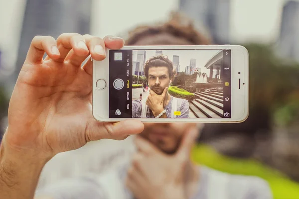 Young attractive man doing selfie phone on the background of skyscrapers. Portrait of a man with intricate facial expressions.Guy holding chin with one hand. — Stock Photo, Image