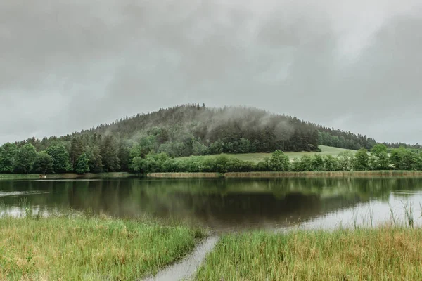 Panoramic view of a lake in front of misty forest. Foggy morning outdoors.Beautiful autumn spring landscape with hill and water reflection.Misty forest scenery.Trees reflected in water.Calm atmosphere