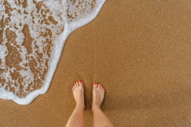 Kumsalda kırmızı tırnaklı mutlu kadın güneşli yaz gününün, dalgaların ve berrak deniz suyunun keyfini çıkarıyor. Çıplak ayaklı kadın bacakları. Seyahat konsepti afişi.