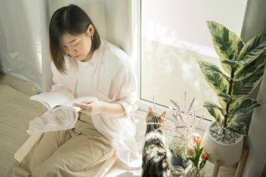 Hala evde Asyalı bir kadının kitap okuması ve kedisiyle oturma odasında oynaması konseptinde.