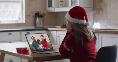 Beyaz kadın evde Noel Baba şapkası takarak, mutfakta oturup arkadaşlarıyla ağır çekimde video sohbeti yapıyor. Noel zamanı, covid 19 salgını sırasında kendini izole etme.