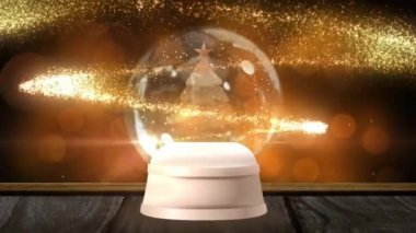 Kayan yıldızların dijital animasyonu, kar küresindeki Noel ağacının etrafında hareket ederken, ahşap yüzeyde, ışığın altın noktalarına karşı. Noel kutlaması geleneği