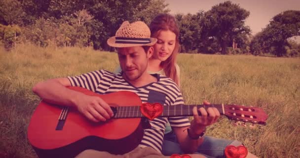 フィールドで女性のためにギターを弾く男の上に浮かぶ赤いハートバルーンのアニメーション ハッピーバレンタインデーのお祝いのコンセプトデジタル生成された画像 — ストック動画