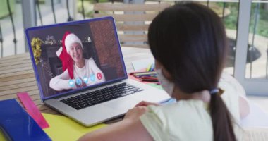 Yüz maskesi takan beyaz bir kız, evde geçirdiği Noel 'de bilgisayarla bir kadınla görüntülü konuşma yapıyor. Covid 19 salgını sırasında aile ve arkadaşlarla iletişim kurmak.