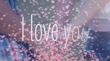 I Love You 'nun animasyonu beyaz harflerle yazılmış, birbirine aşık çiftler el ele tutuşuyor ve arka planda pırıl pırıl pembe kalpler var. Sevgililer Günü kutlaması konsepti dijital bileşik.