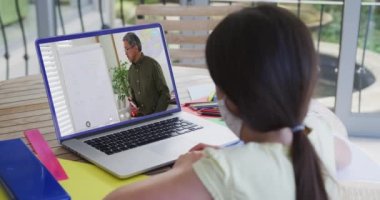 Beyaz maskeli kız, erkek öğretmenle video görüşmesinde dizüstü bilgisayar kullanıyor. Çevrimiçi eğitim karantina süresince evde tek başına kalıyor..