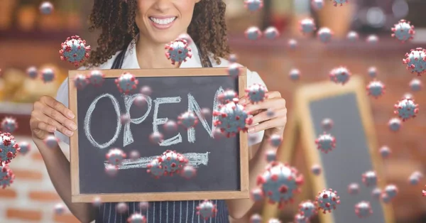 Coronavirus cells floating over mixed race woman chef holding an Open sign, smiling, restaurant in the background. Coronavirus and restaurant business.