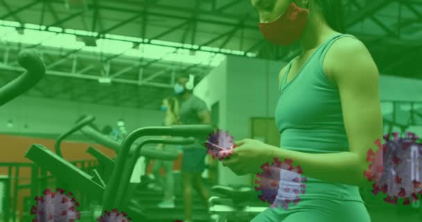 Ковид 19 клеток плавающих против женщины в маске для лица с помощью смартфона в тренажерном зале — стоковое видео