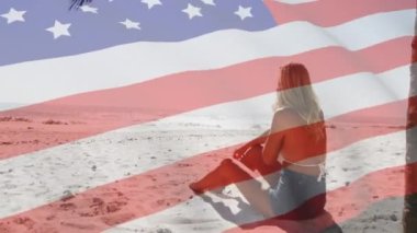 Amerikan bayrağının sahilde akıllı telefondan konuşan bir kadının üzerinde dalgalanması. ABD vatanseverliği ve demokrasi kavramı dijital olarak oluşturulmuş video.