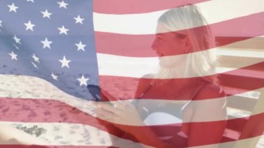 Amerikan bayrağının kumsalda akıllı telefon kullanarak güverte koltuğunda gülümseyen kadını dalgalandırması. ABD vatanseverliği ve demokrasi kavramı dijital olarak oluşturulmuş video.