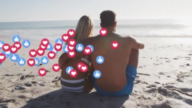 Heart and like icons floating against rear view of caucasian couple sitting on the beach. social media networking and vacation concept