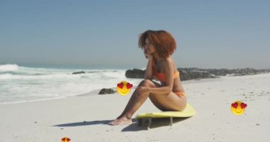 Animation of red heart love emojis digital icons over smiling woman sitting on surfboard on beach. digital interface, social media and global networking concept digitally generated video.