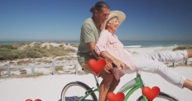 Sahilde bisiklete binen yaşlı çiftin üzerine kalp balonu dijital simgelerinin animasyonu. dijital arayüz, sosyal medya ve küresel teknoloji konsepti dijital olarak oluşturulmuş video.
