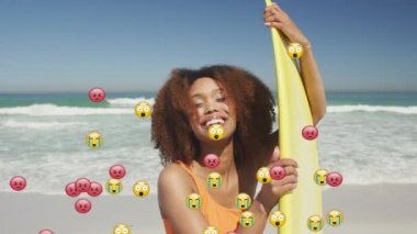 Animation of social media emojis over smiling woman holding surfboard on beach. digital interface, social media and global network concept digitally generated video.