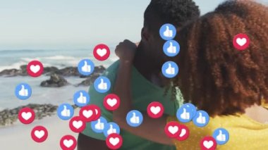 Heart and like icons floating against african american couple embracing each other on the beach. social media networking and vacation concept
