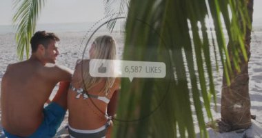 Like icons with increasing numbers against rear view of caucasian couple sitting on the beach. social media networking and vacation concept