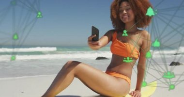 Animation of networks with digital icons over woman taking selfie with smartphone on beach. digital interface, social media and global networking concept digitally generated vide