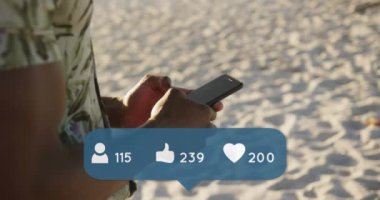 Animation of people, thumbs up and heart icons with numbers over man using smartphone on beach. digital interface, social media and global technology concept digitally generated video.