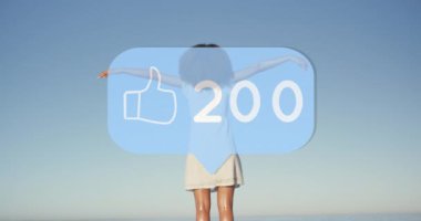 Animation of speech bubble with thumbs up icon and numbers over woman with arms stretched on beach. digital interface, social media and global network concept digitally generated video.