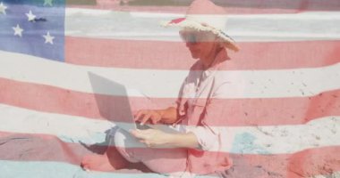 Sahilde otururken dizüstü bilgisayar kullanan yaşlı beyaz bir kadına Amerikan bayrağı sallıyor. Amerika 'nın bağımsızlığı vatansever tatil konsepti