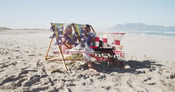 独立日的文字反对资深的高加索夫妇坐在海滩的甲板椅子上 美国独立爱国假日的概念 — 图库视频影像
