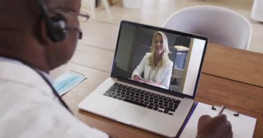 Telefon kulaklığı takan Afrikalı Amerikalı erkek doktor dizüstü bilgisayardan video görüşmesi yaparken not alıyor. uzaktan iletişim ve teletıp danışma kavramı.
