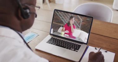 Telefon kulaklığı takan Afrikalı Amerikalı erkek doktor dizüstü bilgisayardan video görüşmesi yaparken not alıyor. uzaktan iletişim ve teletıp danışma kavramı.