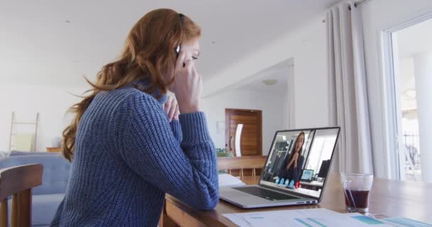 携帯電話のヘッドセットを着用している白人女性は 自宅のラップトップで女性同僚とビデオ通話をしています 家庭の概念から — ストック動画