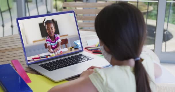 自宅のノートパソコンでアフリカ系アメリカ人の女の子とビデオ通話をしている顔のマスクを身に着けている女の子のバックビュー 遠隔教育とオンライン教育の概念は — ストック動画