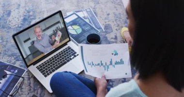 Karışık ırk işkadını dizüstü bilgisayar kullanarak yerde oturuyor ve erkek meslektaşıyla görüntülü konuşma yapıyor. Çevrimiçi toplantı, karantina süresince tecrit altında çalışma.