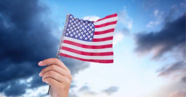 Beyaz elin mavi gökyüzünde bulutlu Amerikan bayrağını tutmasının kompozisyonu. Amerikan vatanseverliği, bağımsızlık ve dijital olarak üretilen görüntü konsepti.