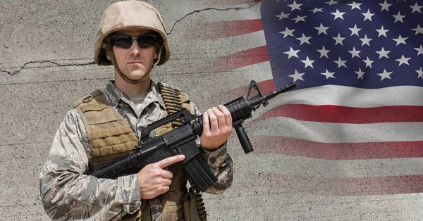 Composition of male soldier wearing sunglasses holding gun, against concrete wall and american flag. patriotism, independence and military concept digitally generated image.