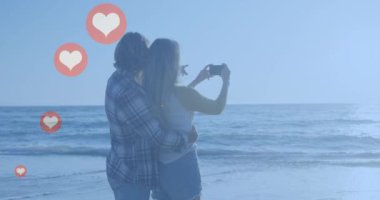 Sahilde fotoğraf çeken beyaz bir çifte karşı birden fazla kırmızı kalp ikonu. Sosyal medya ağı ve teknoloji kavramı