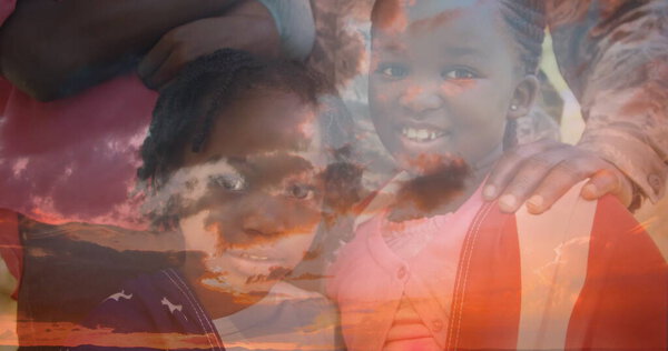 Digital composite of African-American children smiling with an American flag while parents hold their shoulders and background of the sky with clouds