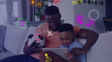 Evdeki kanepede tablet kullanan gülümseyen baba ve oğul üzerine oyun medya simgelerinin animasyonu. küresel iletişim ve video oyunu ağ teknolojisi konsepti dijital olarak oluşturuldu.