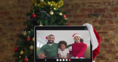Mutlu bir aile, Noel Baba şapkası takıp bilgisayar monitörüne Noel süsleri ve ağaçla görüntülü arama yapıyor. Noel, şenlik ve iletişim teknolojisi dijital kompozit video.