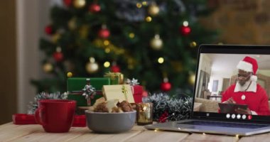 Kıdemli Afro-Amerikalı Noel Baba kostümlü adam dizüstü bilgisayarında Noel ağacıyla görüntülü arıyor. Noel, şenlik ve iletişim teknolojisi dijital kompozit video.