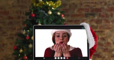 Noel Baba kostümlü beyaz kadın, bilgisayar başında Noel süslemeleri ve ağaçla görüntülü görüşme yapıyor. Noel, şenlik ve iletişim teknolojisi dijital kompozit video.